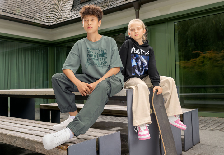 Ein Junge in einem übergroßen grünen T-Shirt und grüner Hose sitzt auf einer Holzbank neben einem Mädchen in einem schwarzen Hoodie und beiger Hose, das ein Skateboard hält.