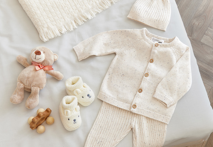 Kuscheliges Baby-Outfit mit Strickjacke, passender Hose, Mütze, Hausschuhen, Teddybär und Holzspielzeug auf einer weichen Kuscheldecke.